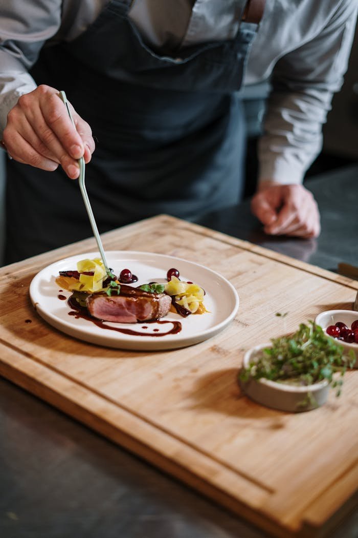 Chef meticulously plates a gourmet dish on a wooden board in a professional kitchen setting.
